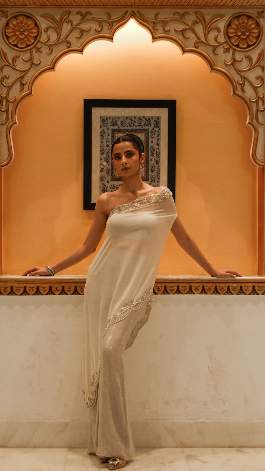 Woman in a white saree standing in front of an ornate wall design.
