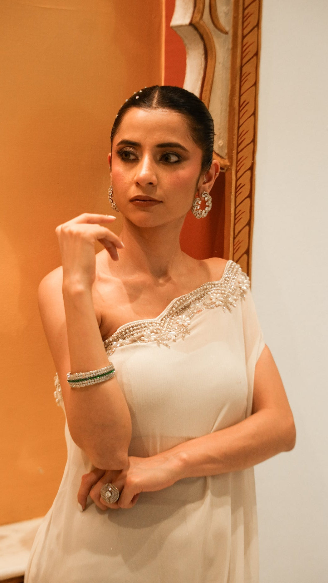 Woman in a white saree with gold detailing standing against an orange wall.