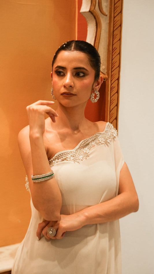 Woman in a white saree with gold detailing standing against an orange wall.