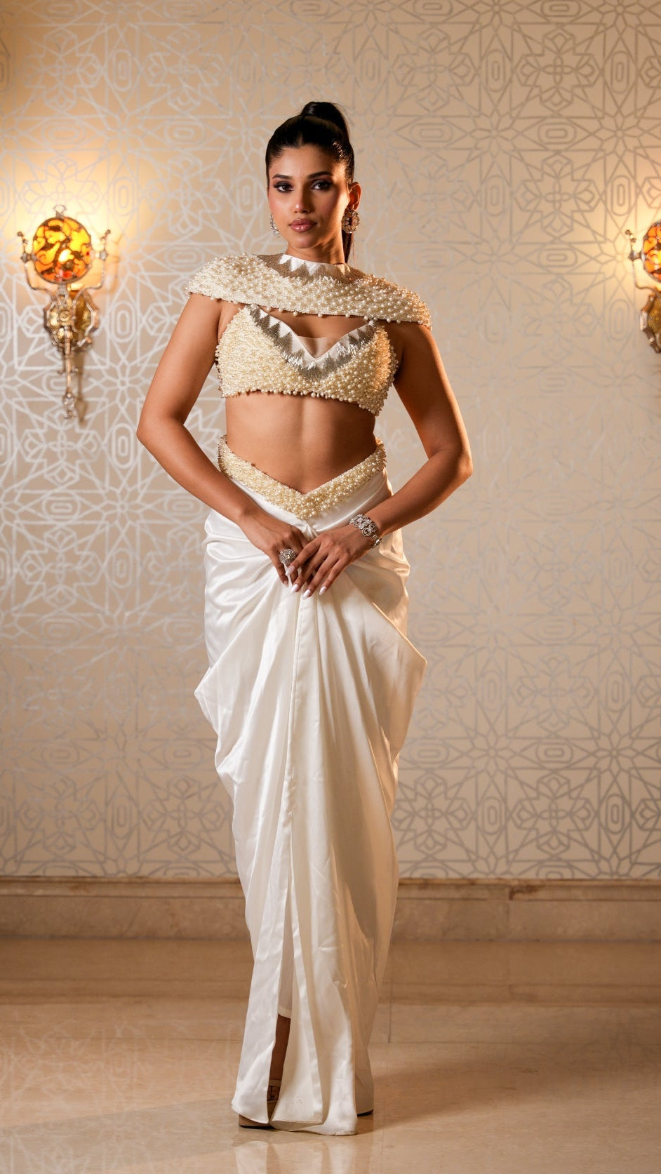 Woman in a traditional white saree with gold jewelry against an ornate wall.