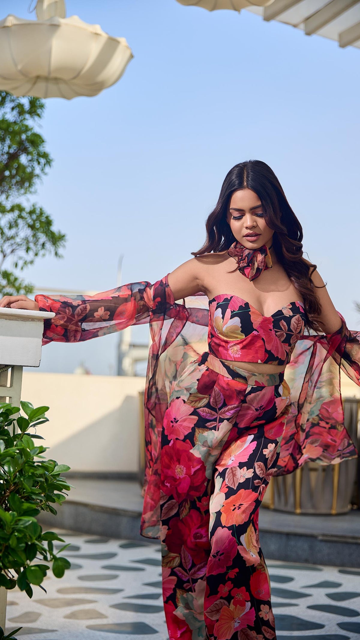 Woman in a floral outfit standing outdoors with plants and a clear sky.