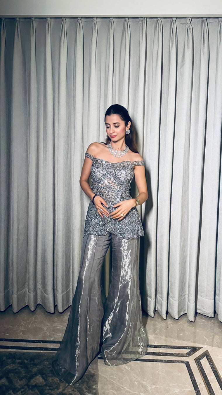Woman in a silver off-shoulder outfit standing in front of a curtain.