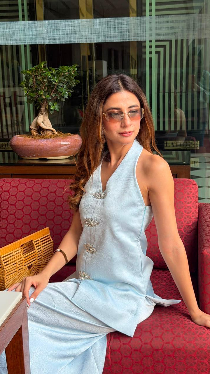 A woman wearing a light blue sleeveless dress with silver embellishments sits on a red patterned sofa in a modern indoor setting.