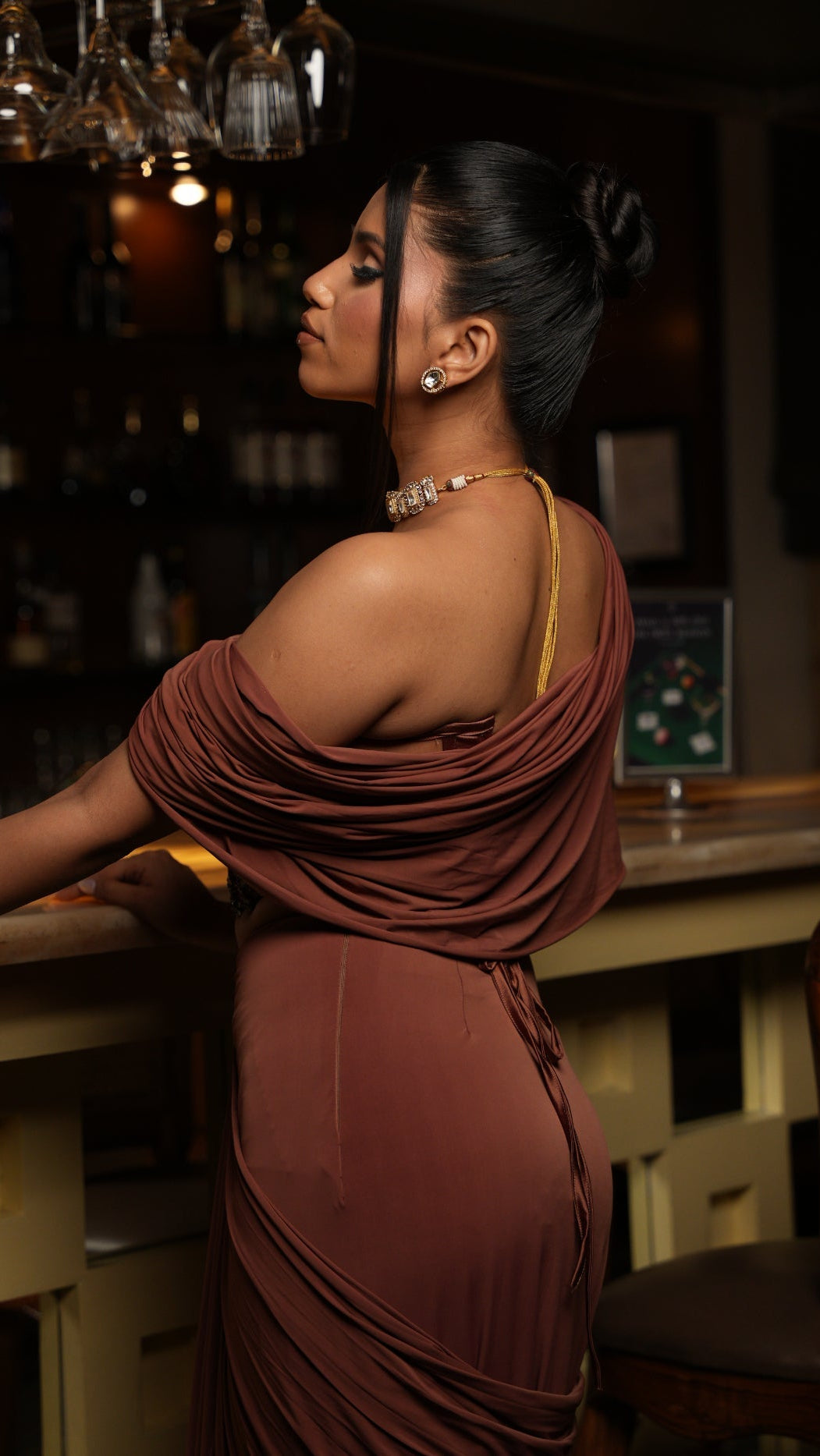 Woman in a brown dress standing at a bar counter with bottles in the background